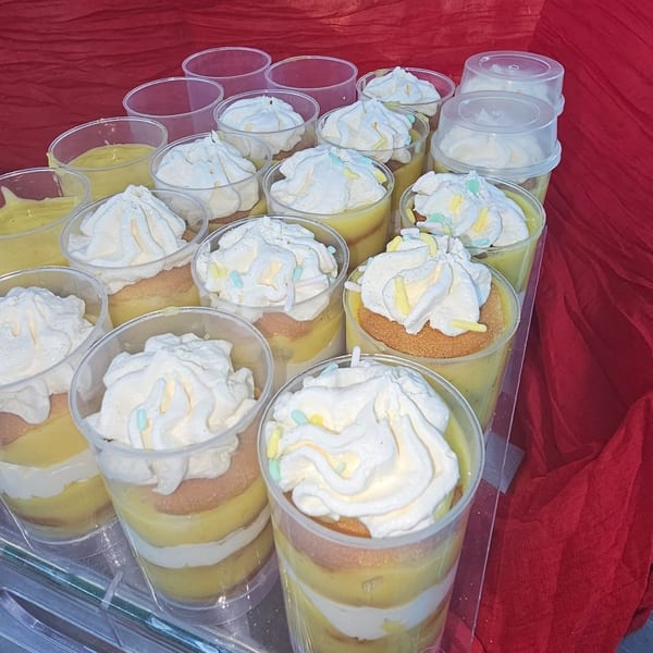 Rows of individual layered desserts in clear cups with whipped cream and sprinkles on a red tablecloth.