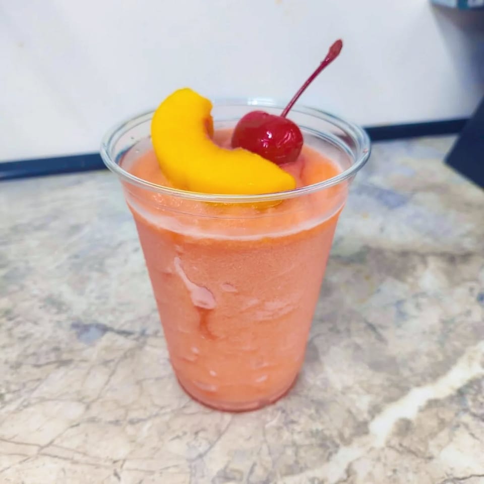 A peach-colored frozen drink in a clear cup, garnished with a peach slice and a cherry.