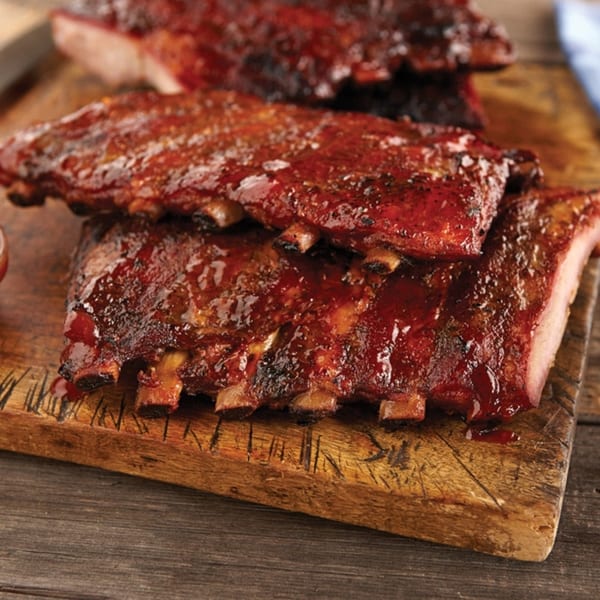 Three racks of glistening BBQ ribs, coated in a rich red sauce, rest on a rustic wooden cutting board.