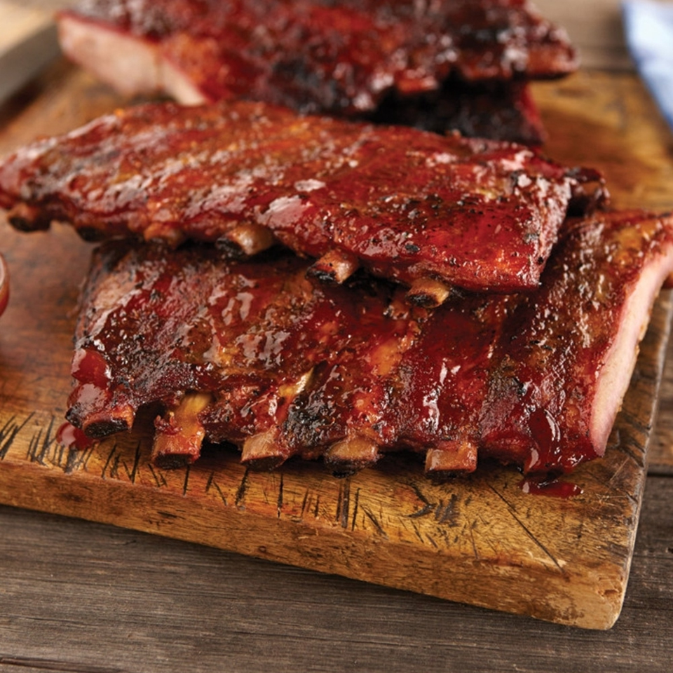 Three racks of glistening BBQ ribs, coated in a rich red sauce, rest on a rustic wooden cutting board.