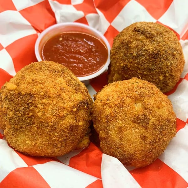 Three golden-brown fried arancini balls with a side of red marinara sauce in a red and white checkered basket.
