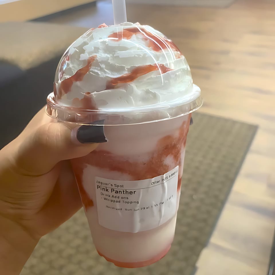 Hand holding a "Pink Panther" drink from Jaguar's Spot, topped with whipped cream and strawberry drizzle.