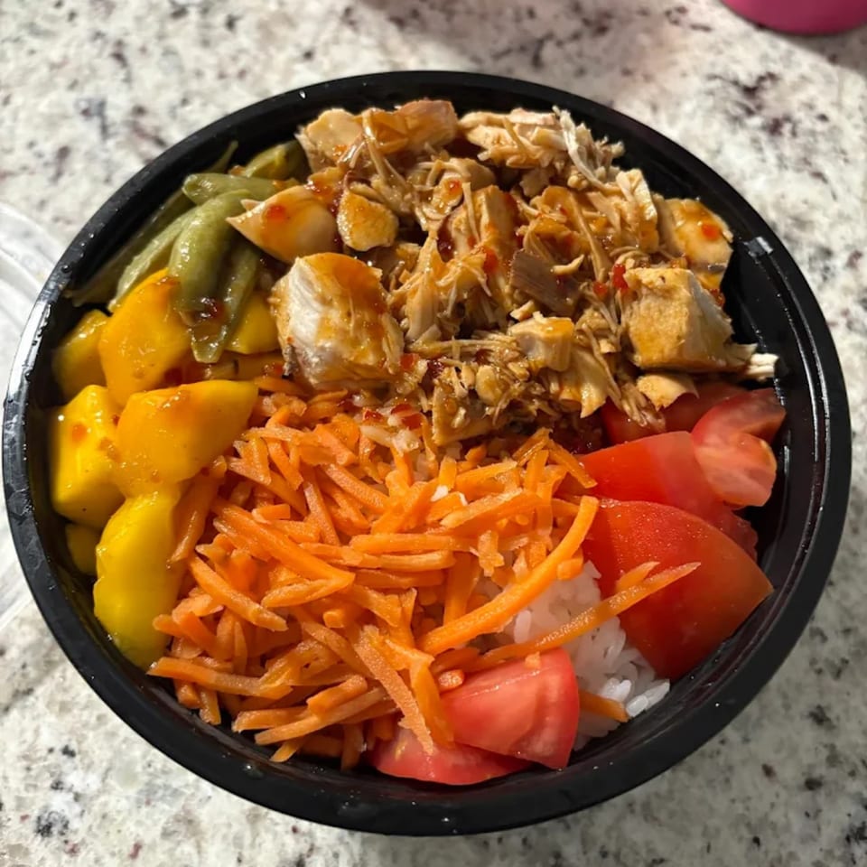 A colorful American bowl with chicken, green beans, mango, shredded carrots, and sliced tomatoes over rice.