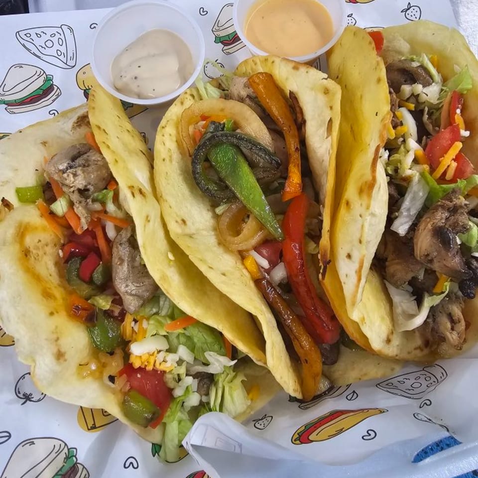 Three soft tacos filled with meat, colorful vegetables, and cheese, with two dipping sauces.