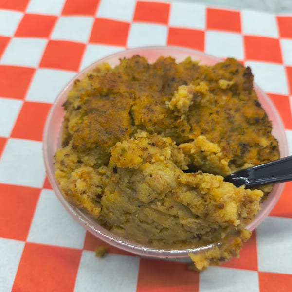 Savory cornbread dressing in a clear plastic bowl with a black spoon, on a red and white checkered tablecloth.