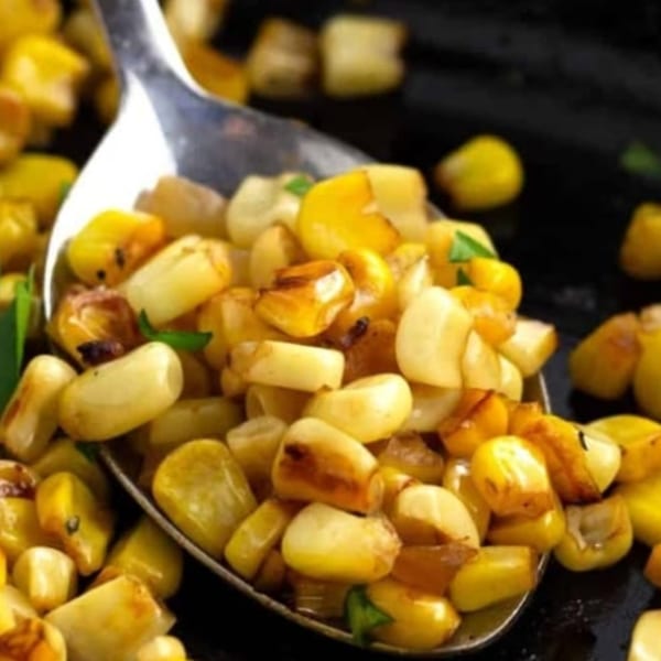 Close-up of a spoon lifting golden-brown roasted corn kernels with green herbs from a dark pan.